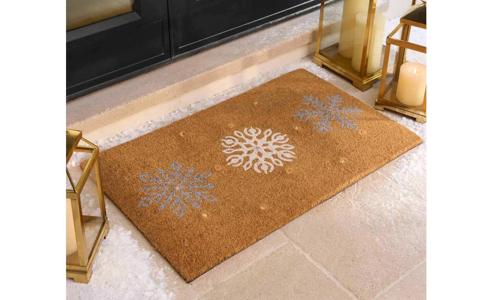 Holiday-themed doormat featuring silver and white snowflake designs on a brown background, placed on a snowy doorstep