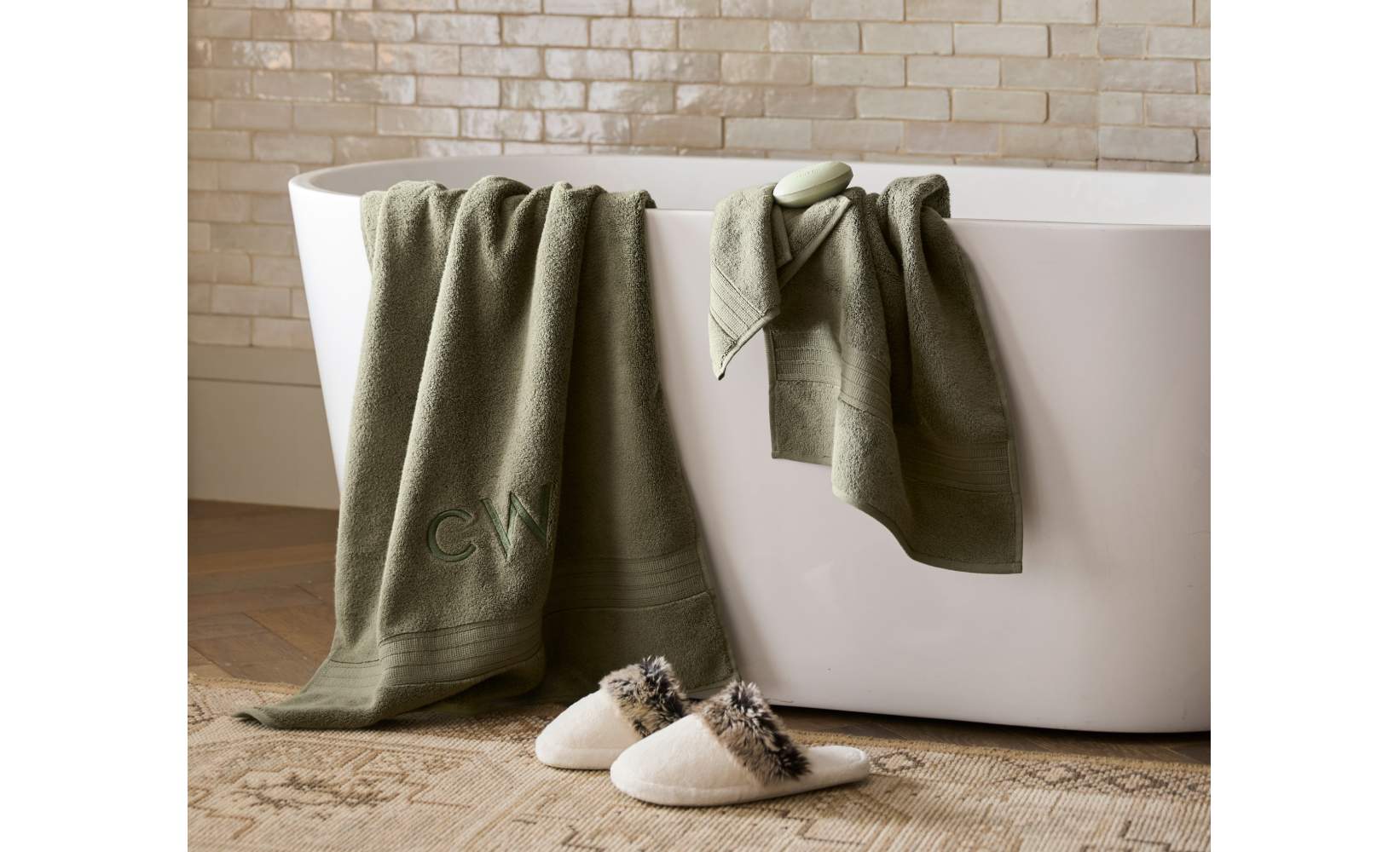 Luxury bathroom decor featuring sage green monogrammed bath towels draped over white freestanding tub, with plush cream slippers on vintage-style rug, creating a spa-like atmosphere Luxury bathroom decor featuring sage green monogrammed bath towels draped over white freestanding tub, with plush cream slippers on vintage-style rug, creating a spa-like atmosphere