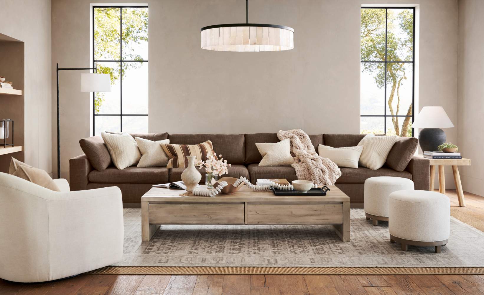 Alt Text: Modern living room with brown sectional sofa, cream swivel chairs and ottomans, featuring large, gridded windows and neutral area rug on hardwood floors.