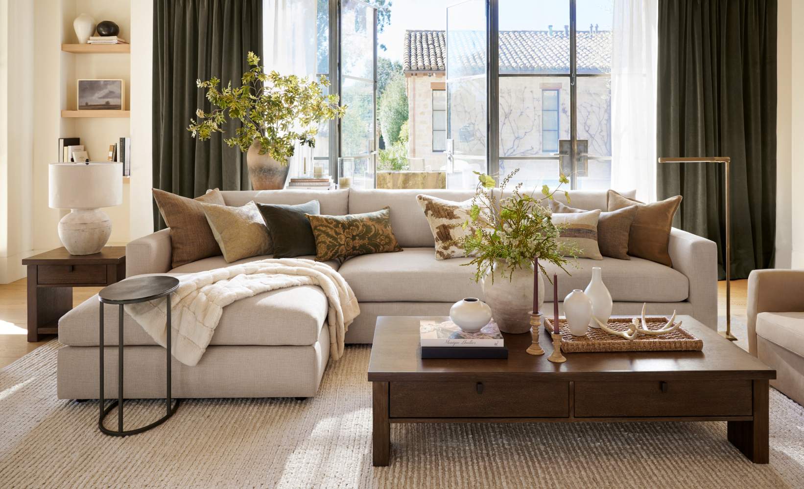 Elegant living room with a beige sectional sofa, earth-toned pillows, wooden coffee table with decorative vases and greenery, and a large window offering natural light. Elegant living room with a beige sectional sofa, earth-toned pillows, wooden coffee table with decorative vases and greenery, and a large window offering natural light.