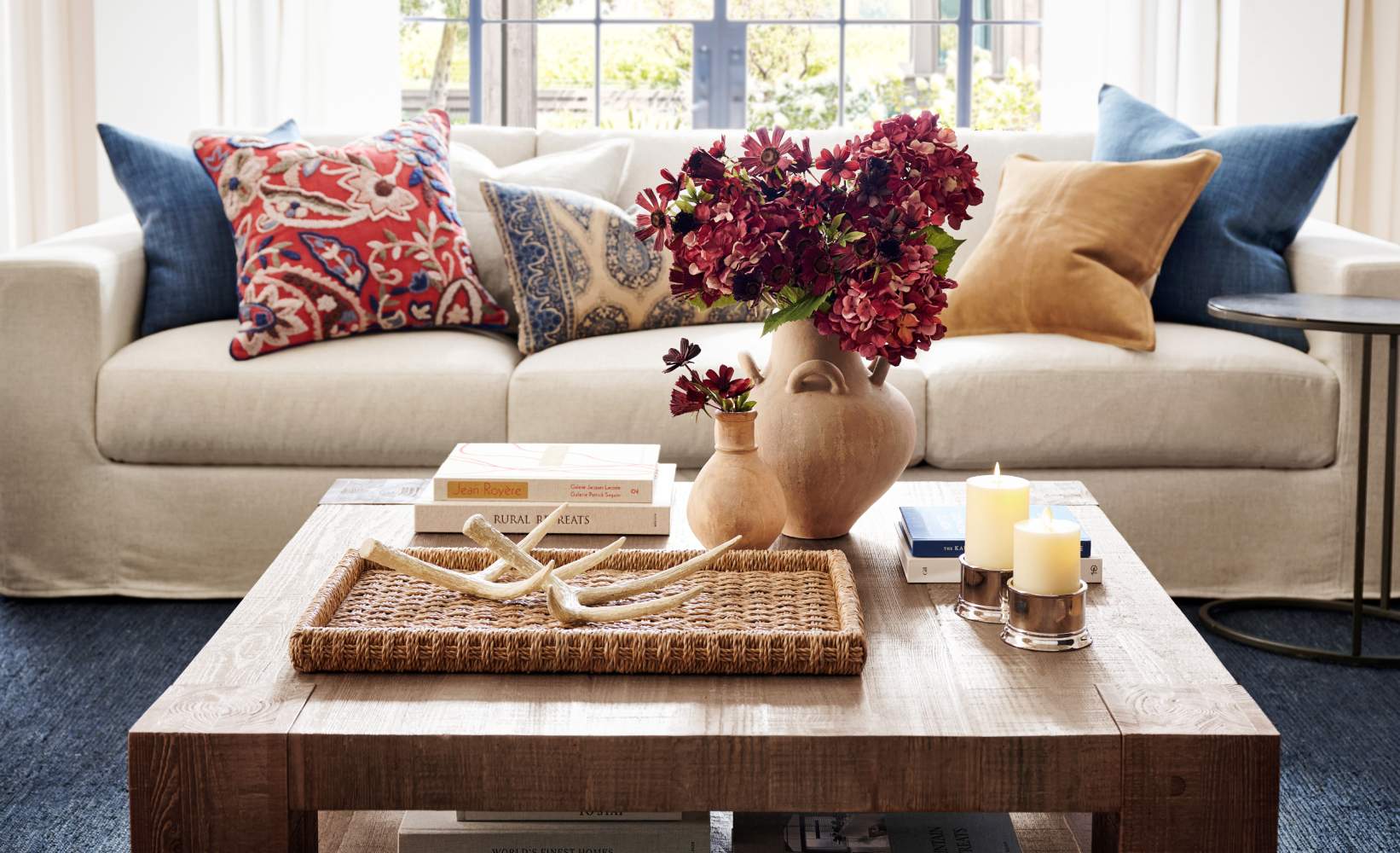 Cozy living room with a beige sofa, colorful patterned throw pillows, wooden coffee table with a woven tray, decorative antlers, candles, and a vase with red flowers. Cozy living room with a beige sofa, colorful patterned throw pillows, wooden coffee table with a woven tray, decorative antlers, candles, and a vase with red flowers.