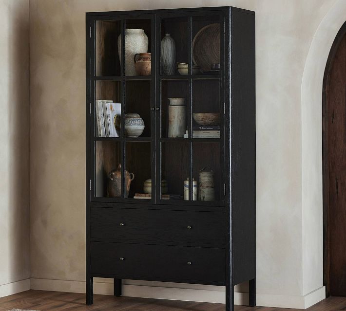 Tall black wood cabinet with glass doors and two bottom drawers, filled with pottery and books.