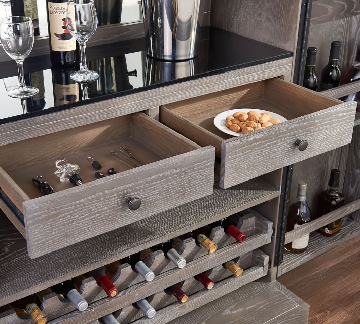 Two open drawers in a wooden cabinet containing bar tools and a plate of snacks with a metal bottle rack.