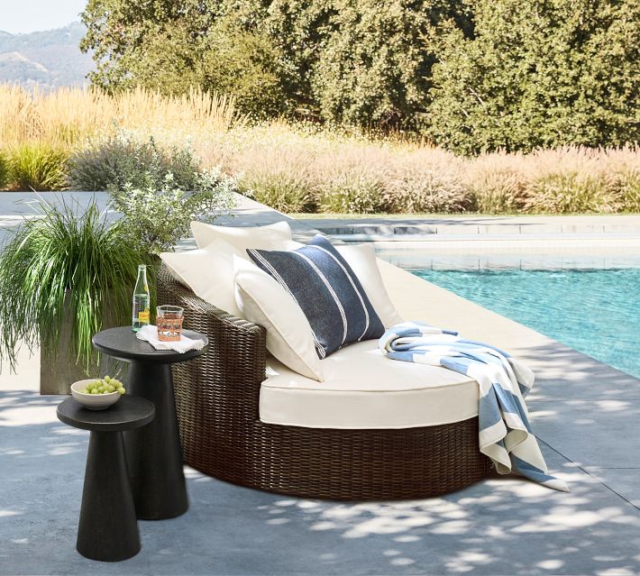 Round brown wicker sofa by pool with beige cushions and blue striped blanket