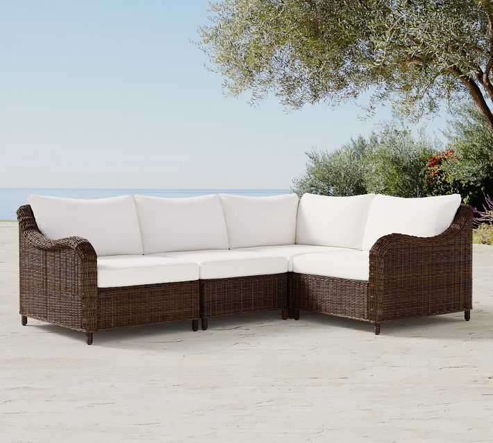 L-shaped brown wicker outdoor sofa set with white cushions on a patio near trees