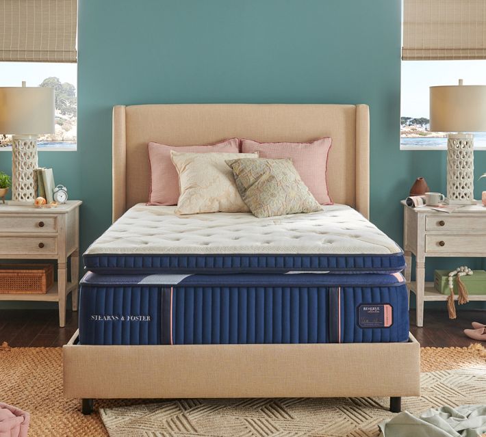 A neatly made bed with a tufted beige headboard and a blue striped mattress shown in a bright, pastel-colored bedroom setting.