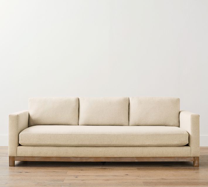 A beige fabric sofa with three back cushions and wooden legs, placed against a white wall