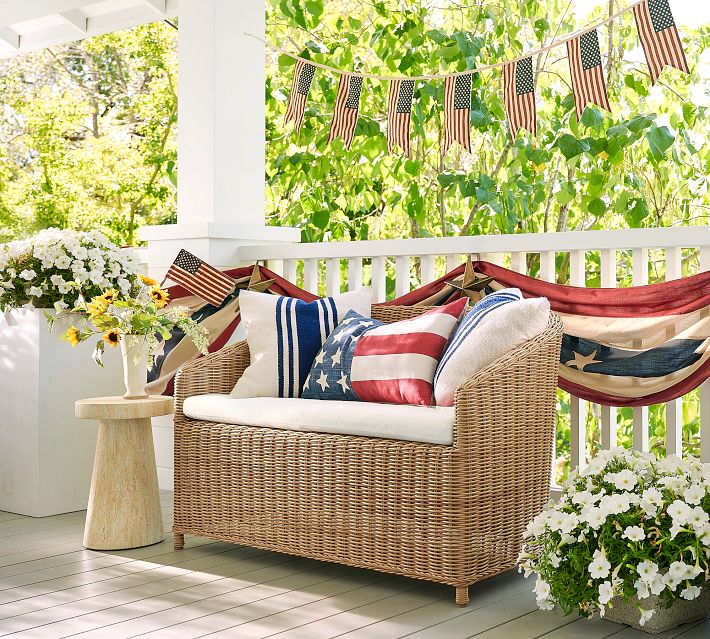 Wicker love-seat with patriotic American flag-style pillows and small table on porch, bunting flags above