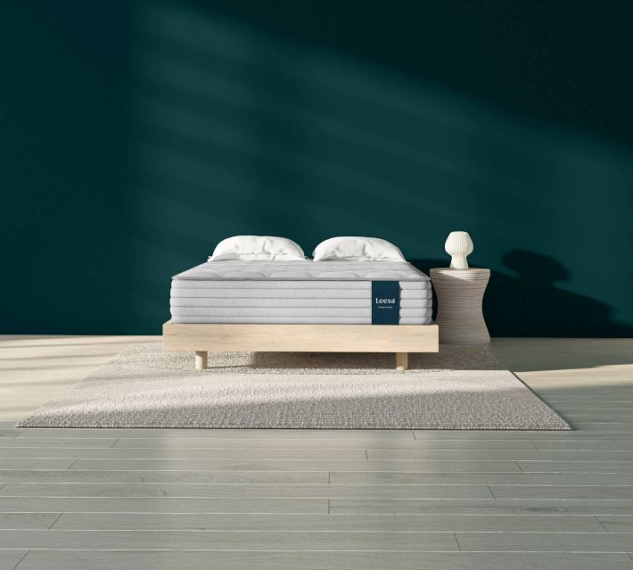 A modern minimalist bed with a thick light gray mattress on a wooden platform, light-colored rug, and teal wall in the background.