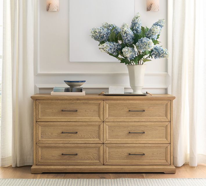 A wooden dresser with six drawers and dark metal handles, decorated with a vase of blue hydrangeas and decor on top.