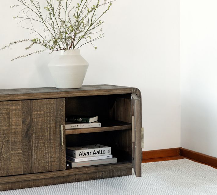 The same dark wooden sideboard with open cabinet revealing shelves containing books, a white vase on top, and a white wall behind.