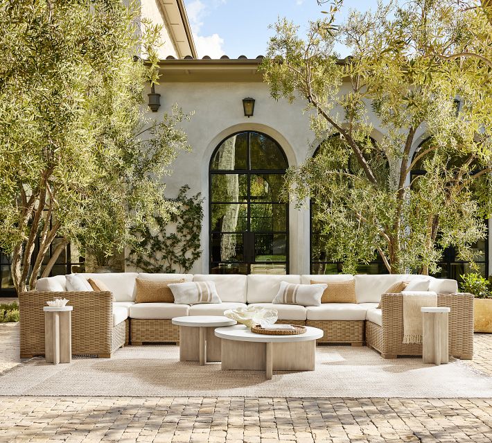 Outdoor living space in front of a villa with a white leather-like sectional sofa set and arched windows framed by trees.