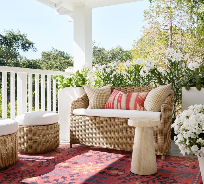 Wicker outdoor loveseat with white seat cushion and striped red/cream pillows, on a porch with greenery