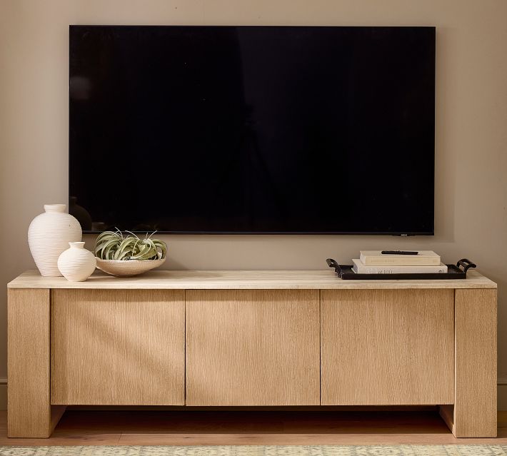 A modern wooden cabinet with a light wood finish supporting a large flat-screen TV, with decorative vases and a tray of books on top