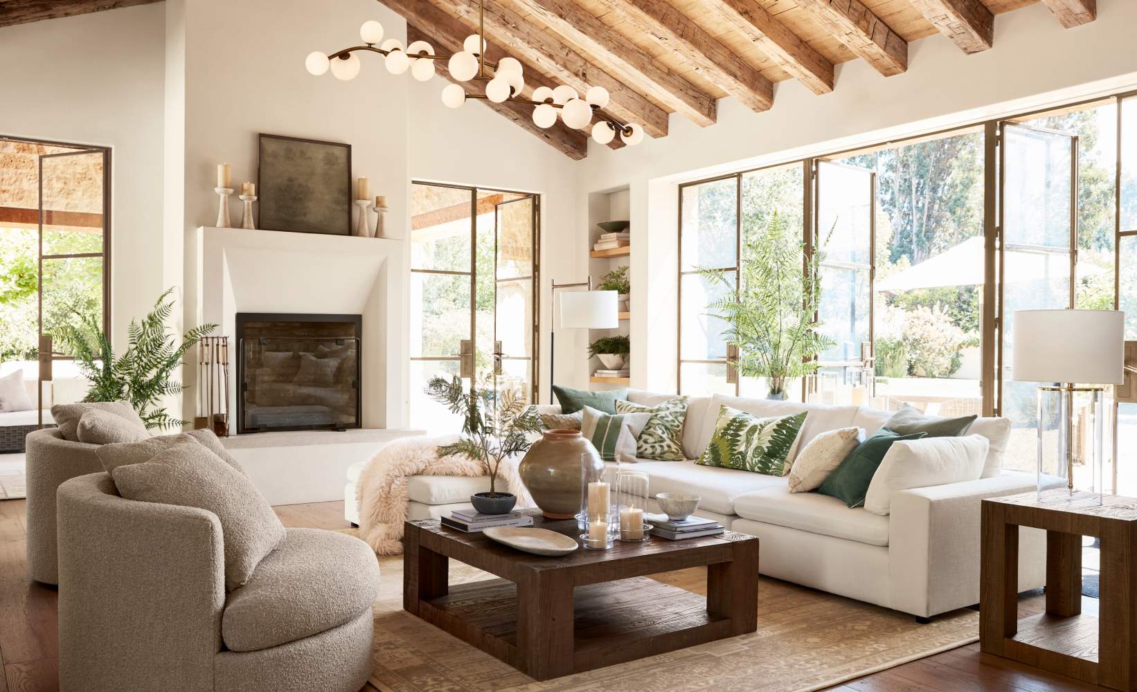 Spacious, light-filled living room with a rustic-modern design featuring a white sectional sofa with neutral throw pillows, two round beige armchairs, and a wooden coffee table.