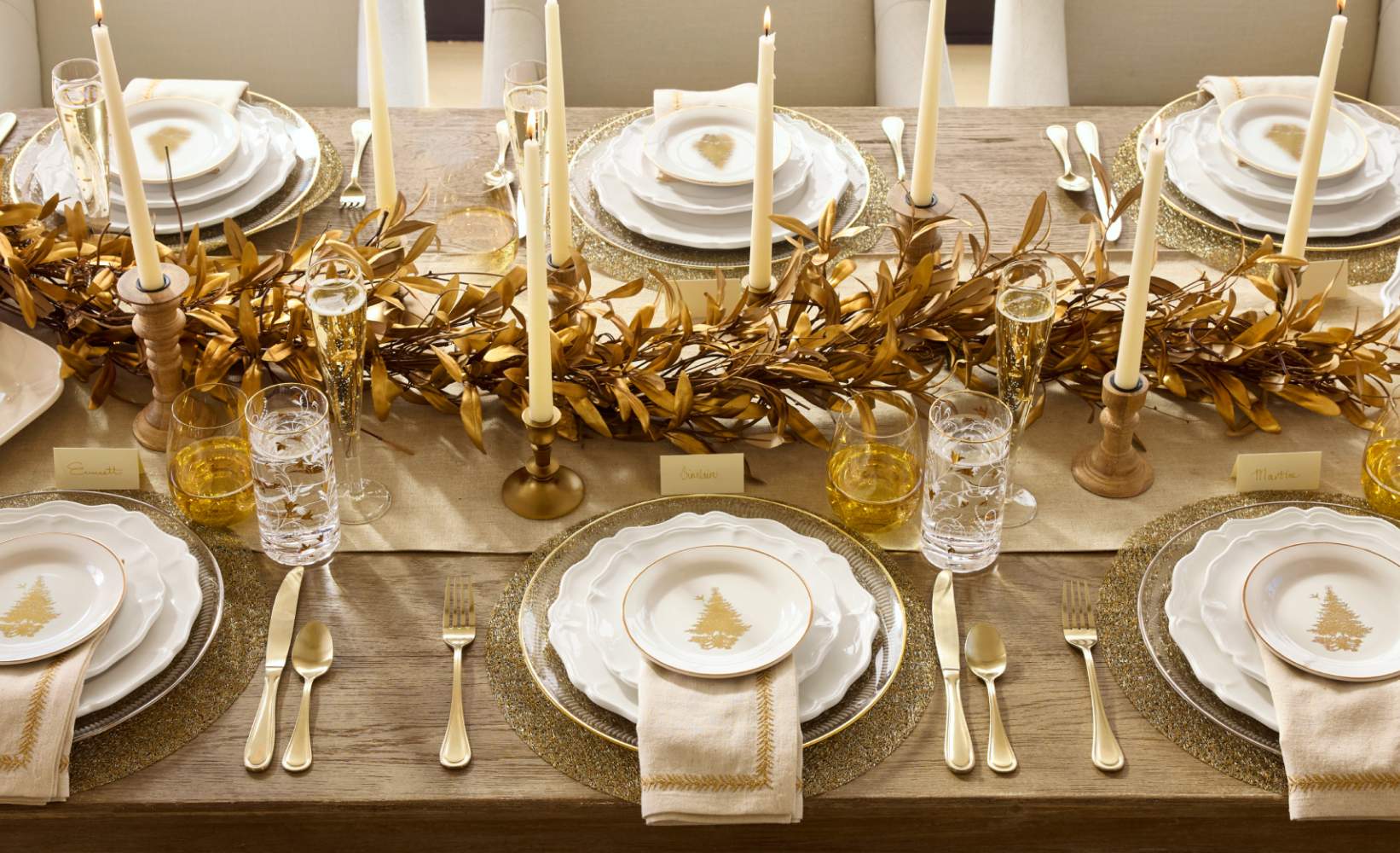 Elegant holiday dining table featuring gold olive leaf garland as a centerpiece, flanked by tall white taper candles in wooden holders.