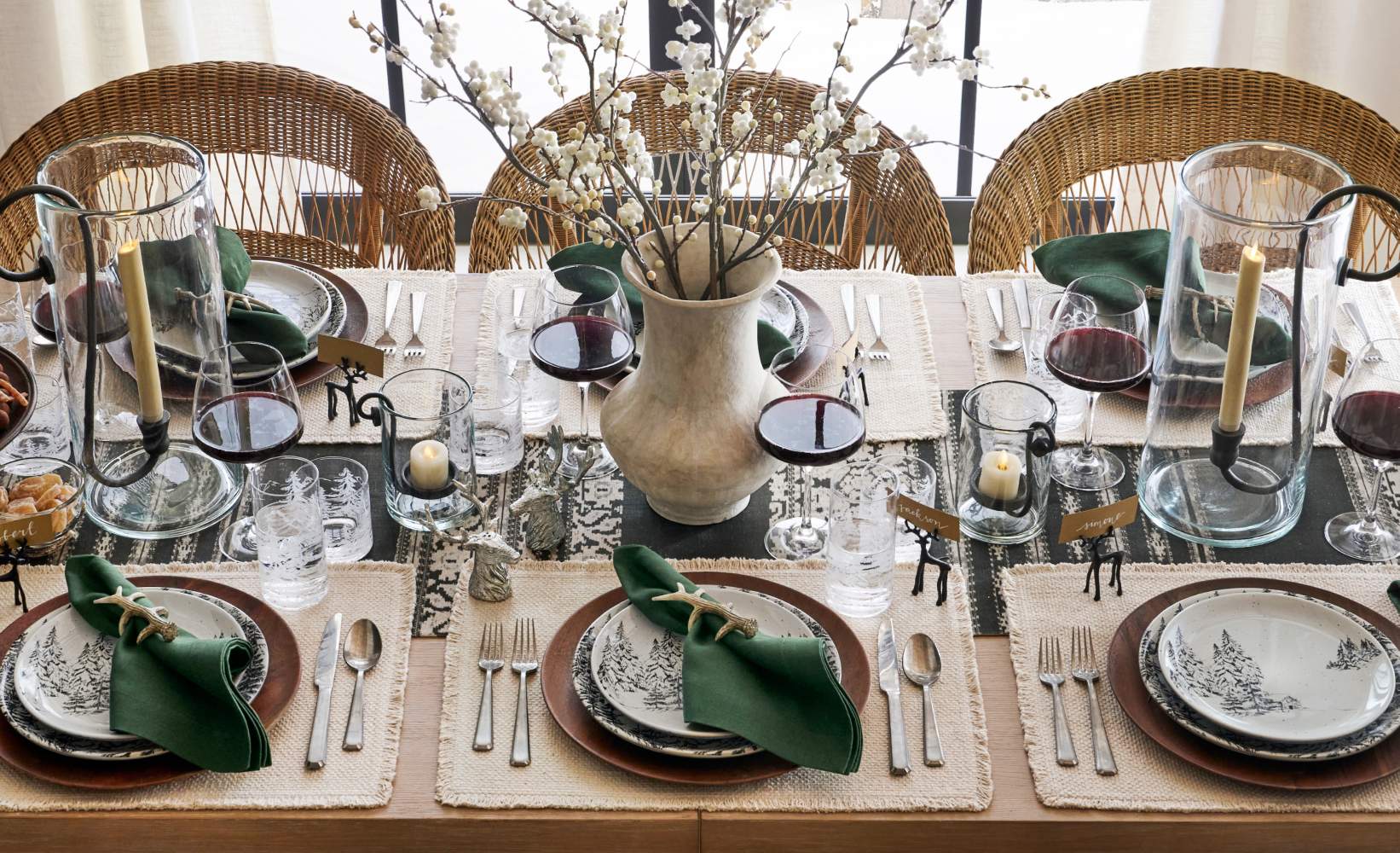Rustic holiday dining table with blooming branches as the centerpiece, surrounded by large glass candle holders with pillar candles.