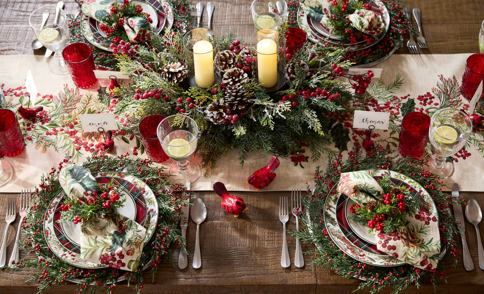 Festive holiday dining table with a Christmas-themed centerpiece featuring evergreen branches, red berries, pinecones, and glowing pillar candles.