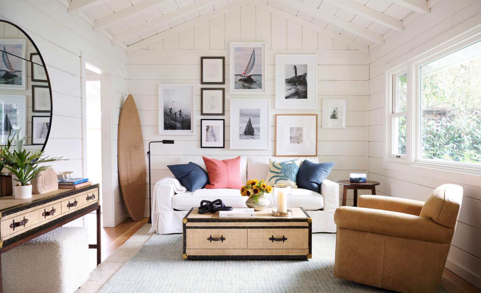 Cozy coastal-style living room with white shiplap walls, framed ocean-themed artwork, surfboard decor, a white sofa with colorful pillows, and rustic storage chest coffee table. Cozy coastal-style living room with white shiplap walls, framed ocean-themed artwork, surfboard decor, a white sofa with colorful pillows, and rustic storage chest coffee table.