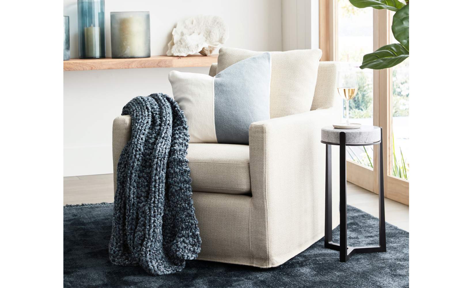 Cozy reading corner featuring cream linen swivel chair with textured throw pillows in white and light blue, chunky knit navy throw blanket, modern black metal side table, and floating wood shelf with coastal decor on a plush navy area rug. Cozy reading corner featuring cream linen swivel chair with textured throw pillows in white and light blue, chunky knit navy throw blanket, modern black metal side table, and floating wood shelf with coastal decor on a plush navy area rug.