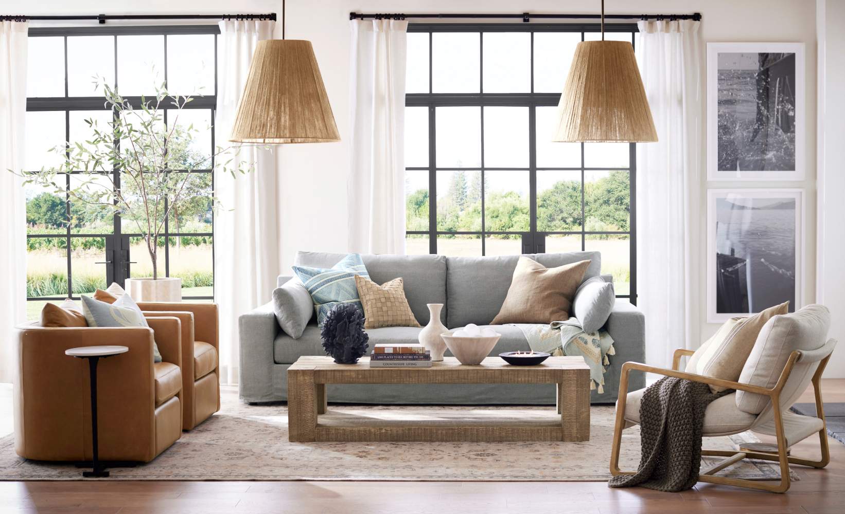 Bright coastal living room with black-framed windows, featuring light gray sofa, tan leather swivel chairs, natural woven pendant lights, modern wood coffee table, and a blonde wood accent chair. Bright coastal living room with black-framed windows, featuring light gray sofa, tan leather swivel chairs, natural woven pendant lights, modern wood coffee table, and a blonde wood accent chair.