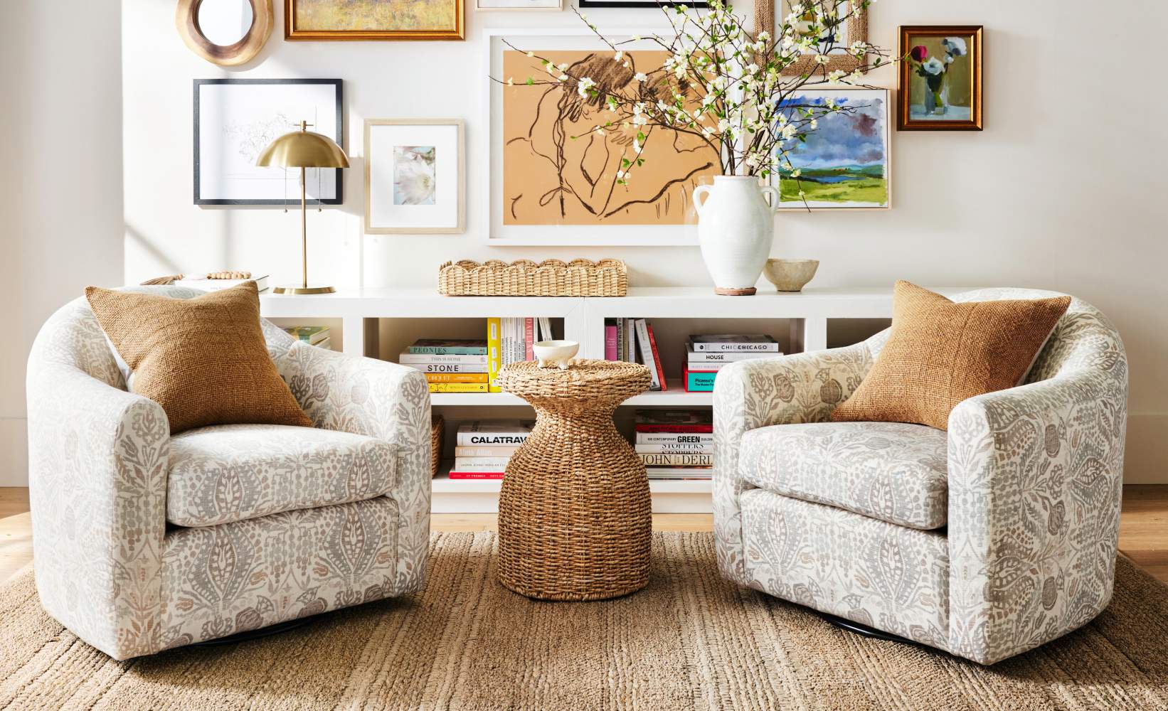 Contemporary reading nook featuring pair of patterned swivel chairs in gray and white botanical print with tan textured pillows, woven rattan hourglass side table, white modern bookshelf, and white ceramic vase with flowering branches on natural jute rug. Contemporary reading nook featuring pair of patterned swivel chairs in gray and white botanical print with tan textured pillows, woven rattan hourglass side table, white modern bookshelf, and white ceramic vase with flowering branches on natural jute rug.