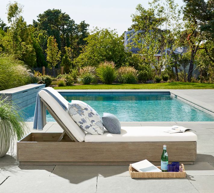 Wooden outdoor daybed with white cushions by pool, decorative pillows, bottle and books nearby