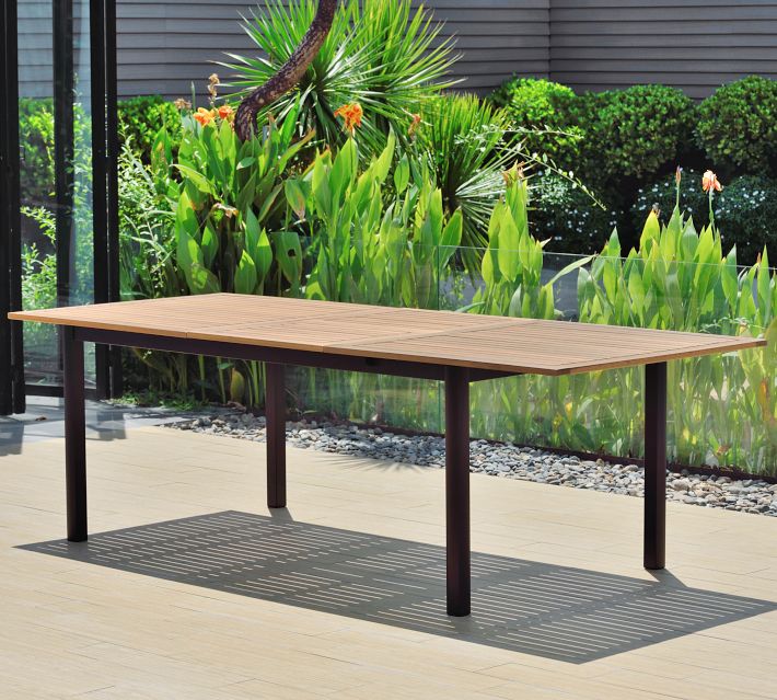 A wooden tabletop with black legs in an outdoor garden setting, plants in background