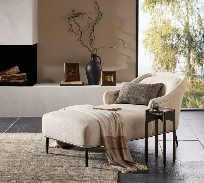 A beige upholstered lounge chair with a matching ottoman in a cozy living room setting, contemporary decor including a vase, books and a throw blanket.