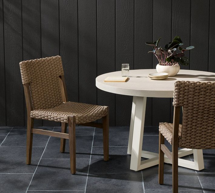 Outdoor round table with woven seating chairs on a dark wall background; dining set in natural tones
