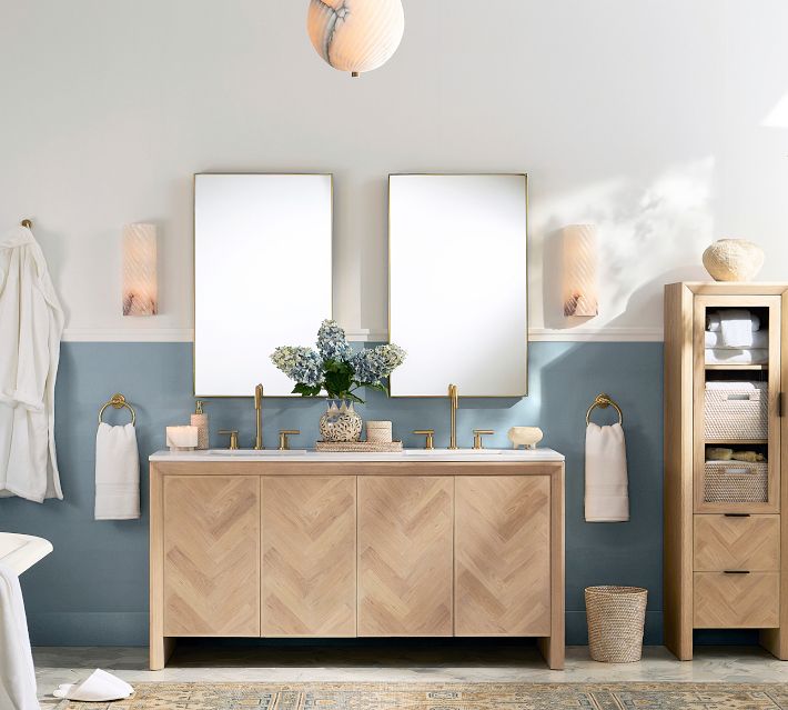 Bathroom vanity with two mirrors and a wooden cabinet base, gold fixtures, and a matching tall cabinet on the right; a vase of flowers on the counter.