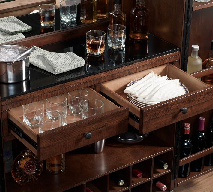 Open drawers showing glassware and a wooden tray with napkins on a dark countertop, with more bottles in background.