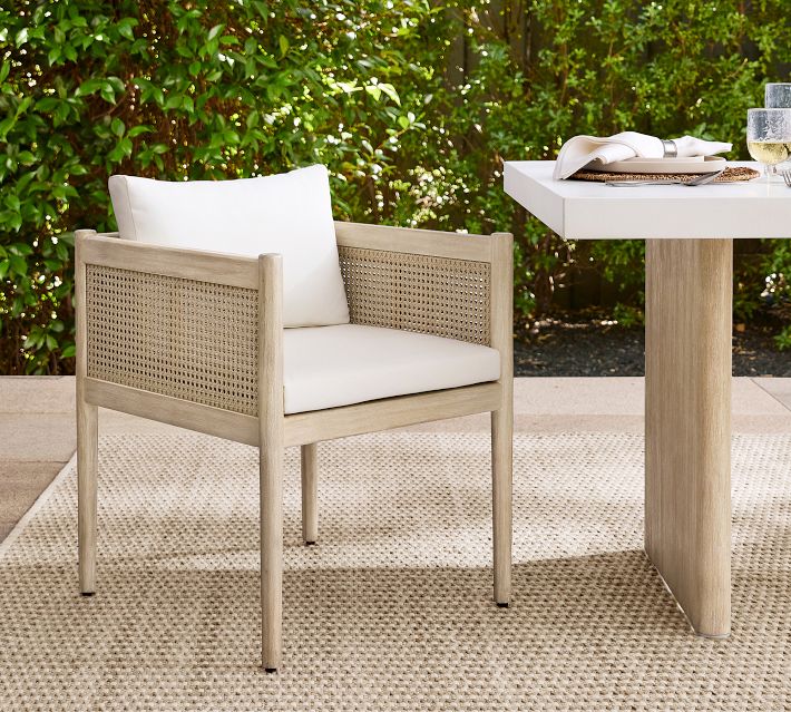 Outdoor dining chair with light wood frame and woven cane side panels, cream cushions, placed on a beige outdoor rug beside a table